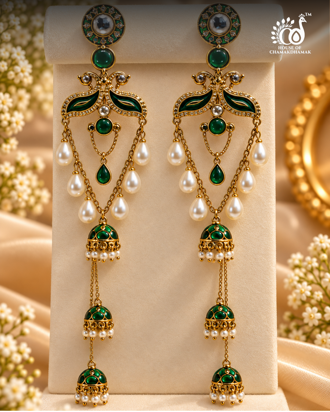 A pair of ornate gold-plated dangle earrings with green enamel, white faux pearls, and clear stones, displayed on a cream jewelry stand with decorative floral props.
