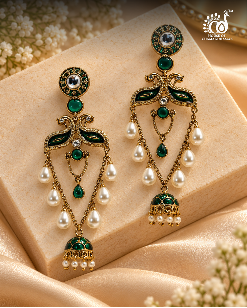 A pair of ornate gold-plated dangle earrings with green enamel, clear stones, and white pearl drops, displayed on a beige stone block with beige satin fabric and small white flowers.